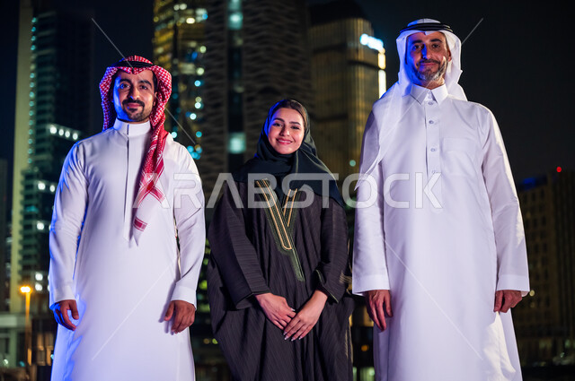 ايماءات السعادة والسرور بالتقدم والنمو, فخر أبناء المملكة بتطورها العمراني، الوقوف باستقامة وثقه واعتزاز بالدولة السعودية والنظر إلى الكاميرا، مواطنون عرب سعوديون مبتسمون يرتدون الزي التقليدي أمام أبراج وناطحات السحاب المضيئة ليلا في مدينة الرياض