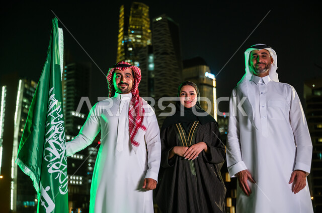 الوقوف باستقامة وفخر بجانب علم المملكة السعودية، العيد الوطني السعودي 8 ربيع الأول، يوم العلم السعودي 11 مارس، مواطنون عرب سعوديون مبتسمون يرتدون الزي التقليدي أمام أبراج وناطحات السحاب المضيئة ليلا في مدينة الرياض، ذكرى اليوم الوطني السعودي