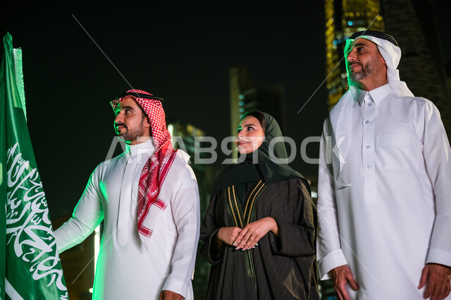 العيد الوطني السعودي، يوم العلم السعودي 11 مارس، مواطنون عرب سعوديون مبتسمون يرتدون الزي التقليدي أمام أبراج وناطحات السحاب المضيئة ليلا في مدينة الرياض، الوقوف بجانب علم المملكة السعودية، ذكرى اليوم الوطني السعودي 8 ربيع الأول ، مناسبات وأعياد وطنية