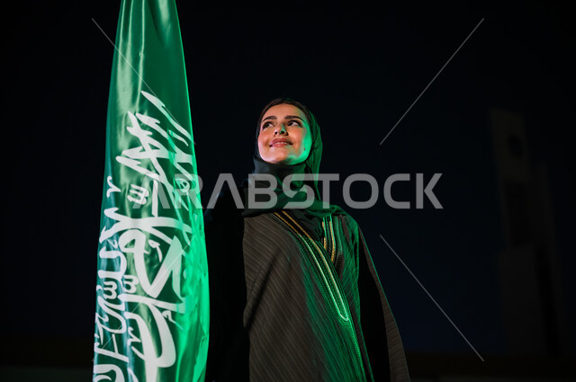 استذكار اليوم الوطني في 23 سبتمبر, ايماءات السعادة والسرور بيوم العلم 11 مارس, صورة مقربة لإمرأة عربية سعودية خليجية مبتسمة ترتدي العباءة والحجاب تمسك علم المملكة باحترام وتقدير, التأمل والنظر إلى شيء ما، مفهوم حب الوطن والانتماء إليه