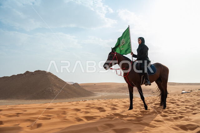 ترسيخ جذور الانتماء الوطني, مشاركة الأحصنة بالمناسبات و الأعياد الوطنية, سيدة عربية خليجية سعودية محجبة تمتطي الخيل وترفع بيدها علم المملكة في الصحراء, الاحتفال بذكرى اليوم الوطني 23 سبتمبر, يوم العلم السعودي 11 مارس, ارتباط الجياد بإرث المملكة