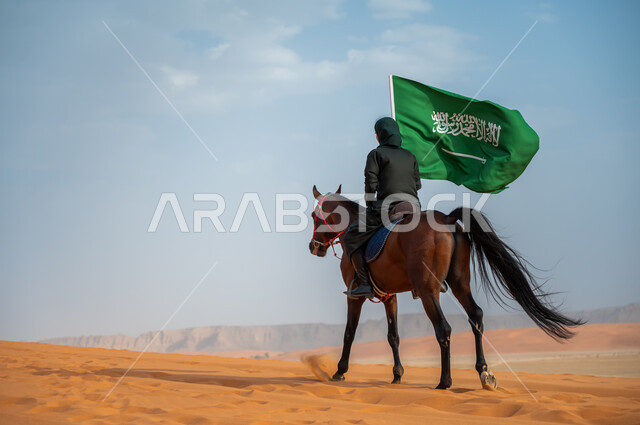 تعميق الانتماء للوطن بالاحتفال بذكرى اليوم الوطني 23 سبتمبر, مشاركة الأحصنة بالمناسبات و الأعياد الوطنية, صورة من الخلف لشابة عربية خليجية سعودية محجبة تمتطي الخيل وترفع بيدها علم المملكة في الصحراء, يوم العلم السعودي 11 مارس