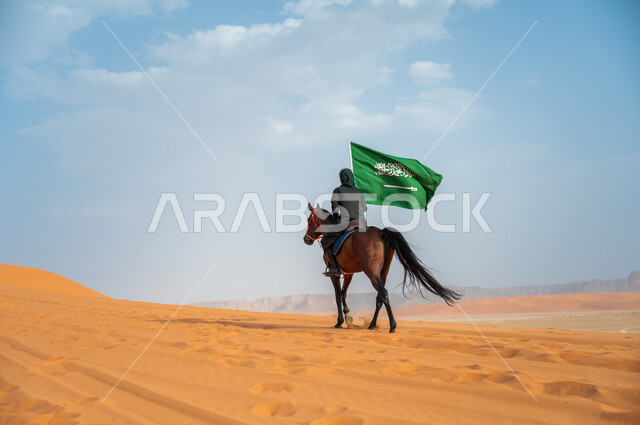 تعميق الانتماء للوطن بالاحتفال بذكرى اليوم الوطني 23 سبتمبر, مشاركة الأحصنة بالمناسبات و الأعياد الوطنية, صورة من الخلف لشابة عربية خليجية سعودية محجبة تمتطي الخيل وترفع بيدها علم المملكة في الصحراء, يوم العلم السعودي 11 مارس