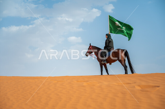 حضارة المملكة وارتباطها بالجياد, مشاركة الأحصنة بالمناسبات الوطنية, صورة من الجانب لفارسة عربية خليجية سعودية محجبة تمتطي الخيل وترفع علم المملكة في الصحراء, تعميق الانتماء للوطن بالاحتفال بذكرى اليوم الوطني 23سبتمبر, يوم العلم السعودي 11 مارس