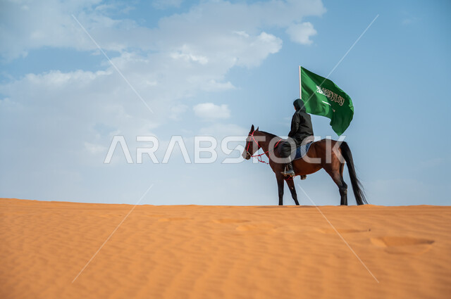 حضارة المملكة وارتباطها بالجياد, مشاركة الأحصنة بالمناسبات الوطنية, صورة من الجانب لفارسة عربية خليجية سعودية محجبة تمتطي الخيل وترفع علم المملكة في الصحراء, تعميق الانتماء للوطن بالاحتفال بذكرى اليوم الوطني 23سبتمبر, يوم العلم السعودي 11 مارس