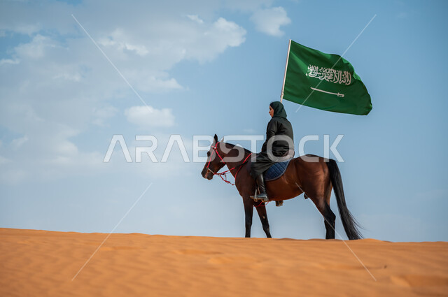 ارتباط الجياد بإرث و تاريخ المملكة, مشاركة الأحصنة بالمناسبات الوطنية, صورة من الجانب لفارسة عربية خليجية سعودية محجبة تمتطي الخيل وترفع علم المملكة في الصحراء, تعميق الانتماء للوطن بالاحتفال بذكرى اليوم الوطني 23سبتمبر, يوم العلم السعودي 11 مارس
