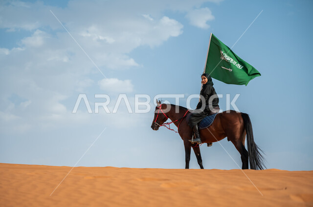 ارتباط الجياد بإرث و تاريخ المملكة, مشاركة الأحصنة بالمناسبات الوطنية, صورة من الجانب لفارسة عربية خليجية سعودية محجبة تمتطي الخيل وترفع علم المملكة في الصحراء, تعميق الانتماء للوطن بالاحتفال بذكرى اليوم الوطني 23سبتمبر, يوم العلم السعودي 11 مارس