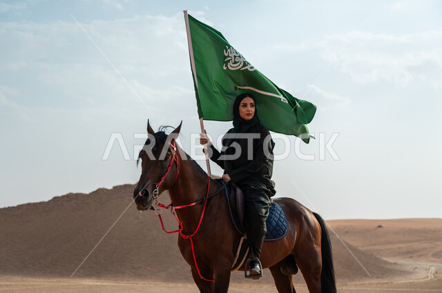 مشاركة الأحصنة بالمناسبات و الأعياد الوطنية, سيدة عربية خليجية سعودية محجبة تمتطي الخيل وترفع بيدها علم المملكة في الصحراء, تعميق الانتماء للوطن بالاحتفال بذكرى اليوم الوطني 23 سبتمبر, يوم العلم السعودي 11 مارس, ارتباط الجياد بإرث وتاريخ المملكة