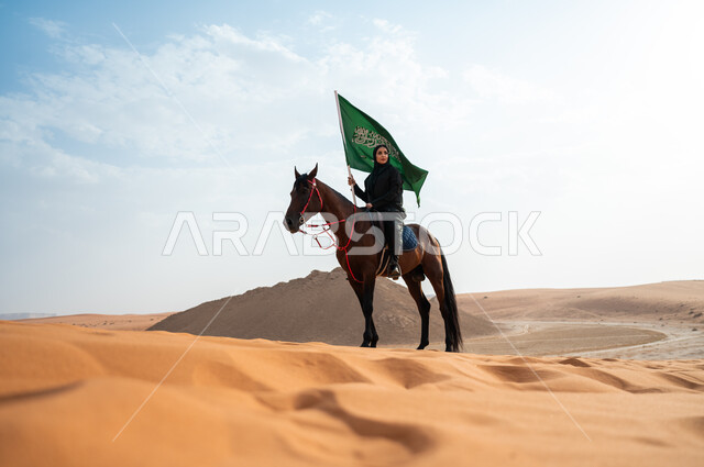 مشاركة الأحصنة بالمناسبات و الأعياد الوطنية, سيدة عربية خليجية سعودية محجبة تمتطي الخيل وترفع بيدها علم المملكة في الصحراء, تعميق الانتماء للوطن بالاحتفال بذكرى اليوم الوطني 23 سبتمبر, يوم العلم السعودي 11 مارس, ارتباط الجياد بإرث وتاريخ المملكة