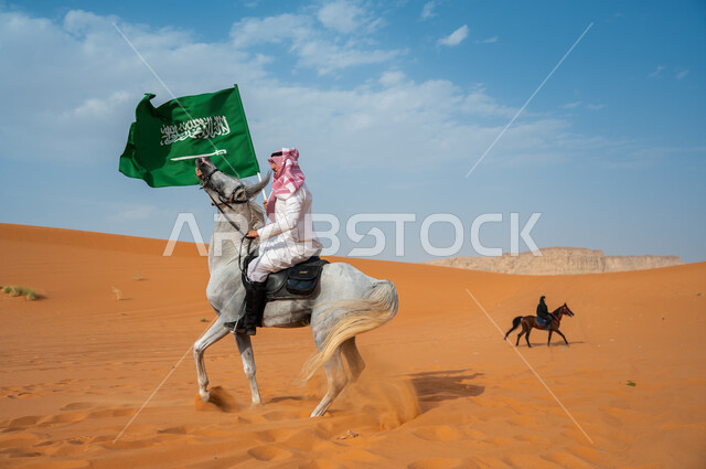 تخليد ذكرى توحيد الوطن، صورة من الجانب لخيال عربي خليجي سعودي يمتطي فرس عربي ويرفع علم السعودية، يوم العلم 11 مارس، العيد الوطني السعودي، اليوم الوطني للمملكة 23 سبتمبر، أنشطة وفعاليات ركوب الخيول في الصحراء، توارث شغف الجياد من الأجداد
