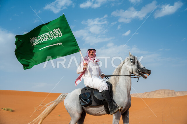تعزيز مفهوم الوحدة الوطنية، صورة من الجانب لرجل عربي خليجي سعودي يمتطي الفرس العربي ويرفع علم السعودية، العيد الوطني 23 سبتمبر، اليوم الوطني لتوحيد المملكة ، يوم العلم السعودي 11 مارس، إظهار مشاعر الاعتزاز بالوطن، هواية ركوب الأحصنة في الطبيعة الصحراوية