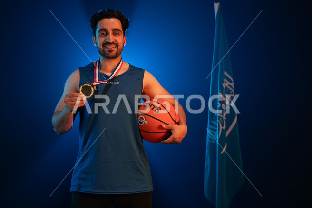 الوقوف بفخر أمام العلم في ذكرى اليوم الوطني في 8 ربيع الأول, بورتريه لمواطن عربي سعودي خليجي مبتسم يرتدي الملابس الرياضية محترف بلعبة كرة السلة بإيماءات السعادة بالفوز بالميدالية الذهبية, يوم العلم 11 مارس، ممارسة هواية اللعب بالباسكيت بول, خلفية زرقاء