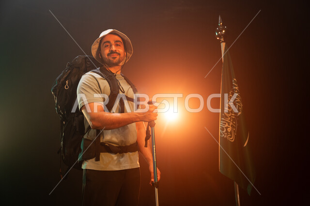 وضع علم السعودية في أعلى القمم الجبلية في ذكرى اليوم الوطني في 23 سبتمبر, الوقوف أمام علم المملكة في يوم العلم 11 مارس, بورتريه لشاب عربي سعودي خليجي مبتسم يحمل حقيبة معدات التسلق, ممارسة هواية الهايكنغ للصحة واللياقة البدنية, خلفية سوداء مع إضاءة ملونة