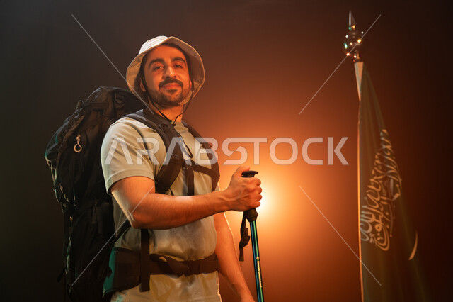الوقوف أمام علم المملكة في يوم العلم 11 مارس, بورتريه لشاب عربي سعودي خليجي مبتسم يحمل حقيبة معدات التسلق, وضع علم السعودية في أعلى القمم الجبلية في ذكرى اليوم الوطني في 23 سبتمبر, ممارسة هواية الهايكنغ للصحة واللياقة البدنية, خلفية سوداء مع إضاءة ملونة