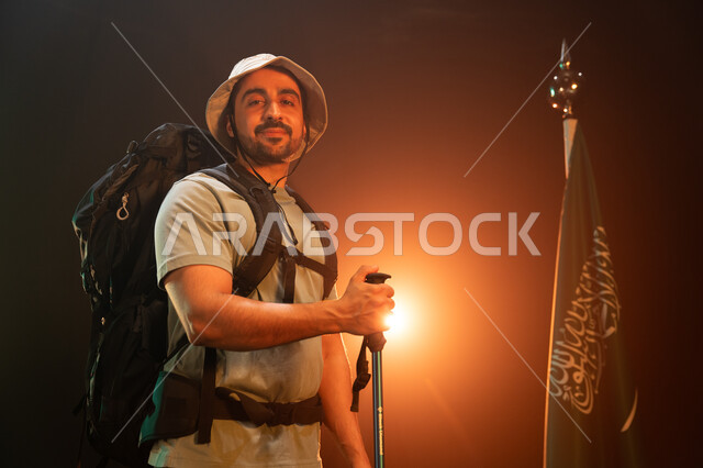 رفع علم السعودية في أعلى القمم الجبلية في ذكرى اليوم الوطني في 23 سبتمبر, الوقوف أمام علم المملكة في يوم العلم 11 مارس، بورتريه لشاب عربي سعودي خليجي يحمل حقيبة معدات التسلق, ممارسة هواية الهايكنغ للصحة واللياقة البدنية, خلفية سوداء مع إضاءة ملونة