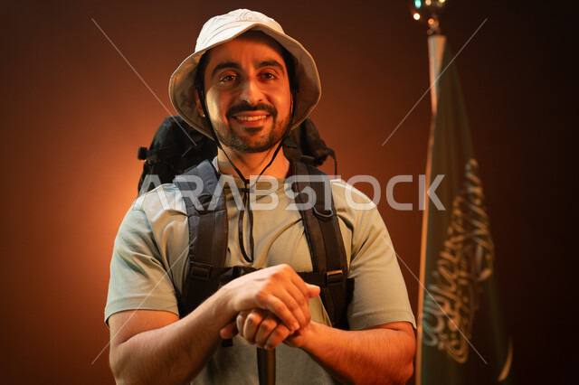 الوقوف بجانب علم المملكة في يوم العلم 11 مارس، بورتريه مقرب لشاب عربي سعودي خليجي مبتسم يحمل حقيبة معدات التسلق, وضع علم السعودية في أعلى القمم الجبلية في ذكرى اليوم الوطني في 23 سبتمبر, ممارسة هواية الهايكنغ اللياقة البدنية, خلفية سوداء مع إضاءة ملونة