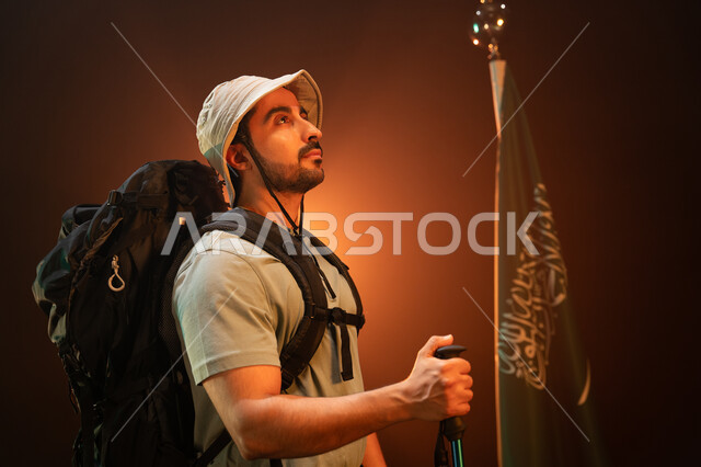 استذكار اليوم الوطني السعودي في 23 سبتمبر, ممارسة رياضة الهايكنغ للصحة واللياقة البدنية, بورتريه مقرب من الجانب لشاب عربي سعودي خليجي يرتدي زي التسلق يقف أمام علم المملكة, النظر إلى شيء ما والمشاركة بالهوايات الترفيهية الممتعه, يوم العلم 11 مارس