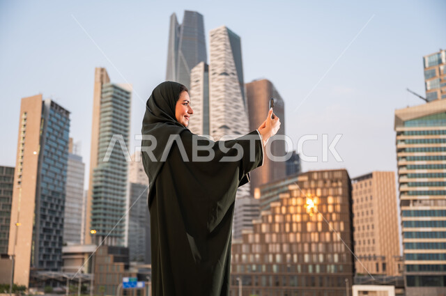 توثيق اللحظات السعيدة عبر الجوال, الأبراج وناطحات السحاب في الرياض في وضح النهار، إمرأة عربية خليجية سعودية ترتدي العباءة والحجاب تقوم بالتقاط صور سيلفي تذكارية باستخدام الهاتف المحمول امام المعالم السياحية، خلفية أبراج مركز الملك عبد الله المالي.