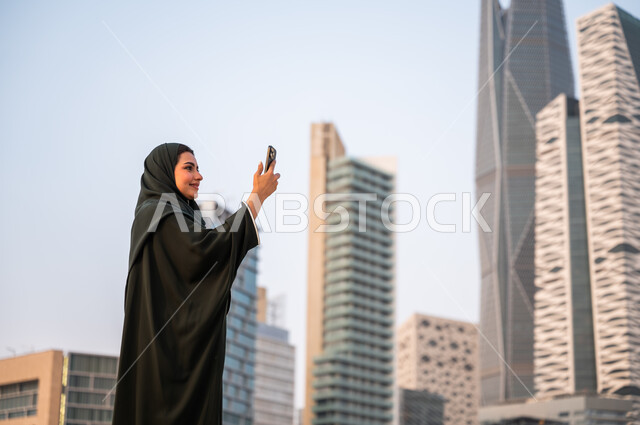 توثيق اللحظات السعيدة عبر الجوال, الأبراج وناطحات السحاب في الرياض في وضح النهار، إمرأة عربية خليجية سعودية ترتدي العباءة والحجاب تقوم بالتقاط صور سيلفي تذكارية باستخدام الهاتف المحمول امام المعالم السياحية، خلفية أبراج مركز الملك عبد الله المالي.