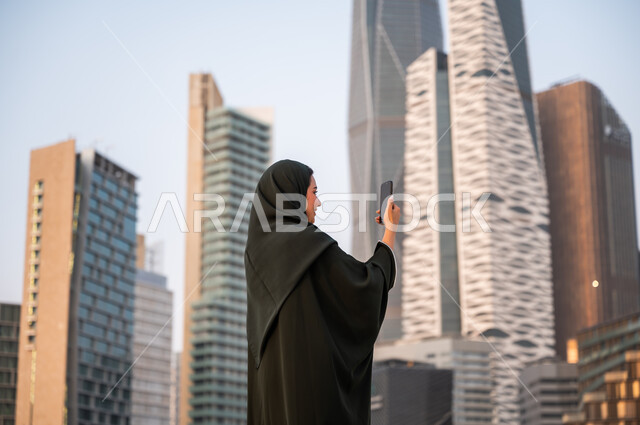 التقاط صور سيلفي تذكارية باستخدام الهاتف المحمول امام المعالم السياحية، الأبراج وناطحات السحاب في الرياض في وضح النهار، إمرأة عربية خليجية سعودية ترتدي العباءة والحجاب تقوم بتوثيق اللحظات السعيدة عبر الجوال, خلفية أبراج مركز الملك عبد الله المالي.