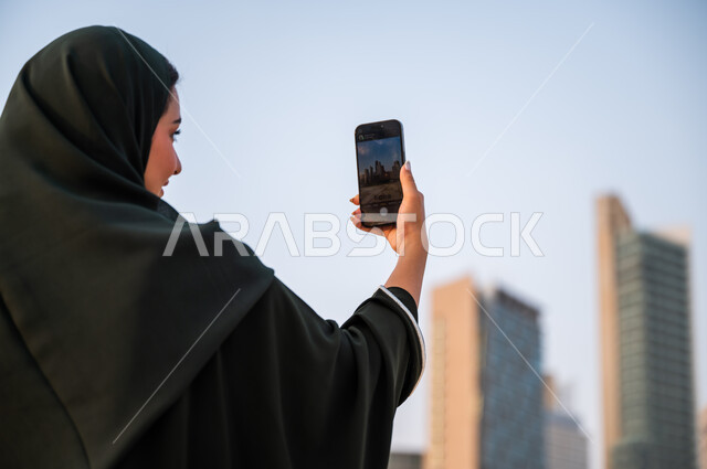 مشاركة ونشر التطور الحضاري والعمراني لمعالم الرياض على وسائل التواصل الاجتماعي، صورة مقربة من الخلف لشابة عربية خليجية سعودية ترتدي العباءة والحجاب تمسك بيدها الهاتف المحمول، التقاط صور تذكارية لأبراج وناطحات سحاب المملكة العربية السعودية