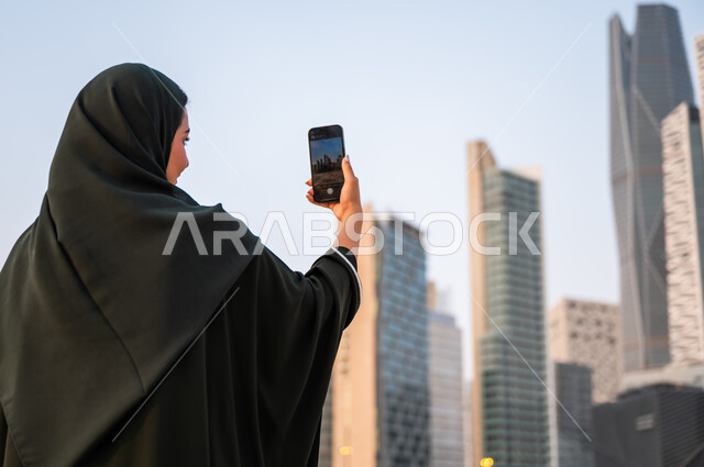 مشاركة ونشر التطور الحضاري والعمراني لمعالم الرياض على وسائل التواصل الاجتماعي، صورة مقربة من الخلف لشابة عربية خليجية سعودية ترتدي العباءة والحجاب تمسك بيدها الهاتف المحمول، التقاط صور تذكارية لأبراج وناطحات سحاب المملكة العربية السعودية