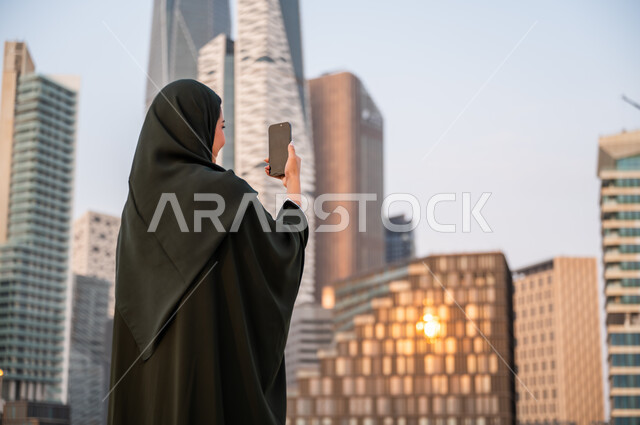نشر ومشاركة التطور الحضاري والعمراني للمنشآت الاقتصادية ومعالم الرياض على وسائل التواصل الاجتماعي، صورة مقربة من الخلف لشابة عربية خليجية سعودية ترتدي العباءة والحجاب تمسك بيدها الهاتف المحمول، التقاط صور تذكارية لأبراج مركز الملك عبدالله المالي