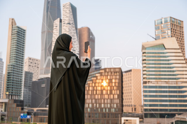 نشر ومشاركة التطور الحضاري والعمراني للمنشآت الاقتصادية ومعالم الرياض على وسائل التواصل الاجتماعي، صورة مقربة من الخلف لشابة عربية خليجية سعودية ترتدي العباءة والحجاب تمسك بيدها الهاتف المحمول، التقاط صور تذكارية لأبراج مركز الملك عبدالله المالي