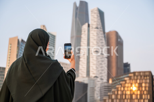 نشر ومشاركة التطور الحضاري والعمراني للمنشآت الاقتصادية ومعالم الرياض على وسائل التواصل الاجتماعي، صورة مقربة من الخلف لشابة عربية خليجية سعودية ترتدي العباءة والحجاب تمسك بيدها الهاتف المحمول، التقاط صور تذكارية لأبراج مركز الملك عبدالله المالي