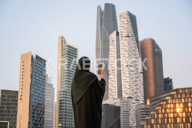 التقاط صور تذكارية لأبراج مركز الملك عبدالله المالي, نشر ومشاركة التطور الحضاري والعمراني للمنشآت الاقتصادية ومعالم الرياض على وسائل التواصل الاجتماعي، صورة من الخلف لشابة عربية خليجية سعودية ترتدي العباءة والحجاب تمسك بيدها الهاتف المحمول