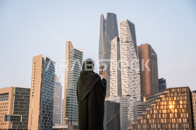 التقاط صور تذكارية لأبراج مركز الملك عبدالله المالي, نشر ومشاركة التطور الحضاري والعمراني للمنشآت الاقتصادية ومعالم الرياض على وسائل التواصل الاجتماعي، صورة من الخلف لشابة عربية خليجية سعودية ترتدي العباءة والحجاب تمسك بيدها الهاتف المحمول