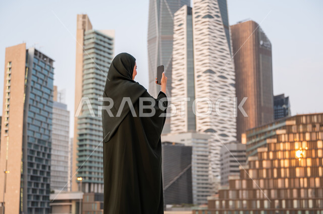 التقاط صور تذكارية لأبراج مركز الملك عبدالله المالي, نشر ومشاركة التطور الحضاري والعمراني للمنشآت الاقتصادية ومعالم الرياض على وسائل التواصل الاجتماعي، صورة من الخلف لشابة عربية خليجية سعودية ترتدي العباءة والحجاب تمسك بيدها الهاتف المحمول