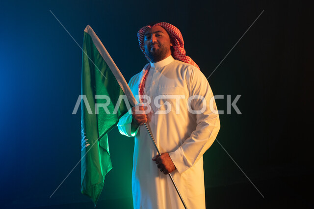 إظهار مشاعر الاعتزاز بالوطن, استذكار اليوم الوطني في 23 سبتمبر, بورتريه لشاب عربي سعودي خليجي يرتدي الشماغ السعودي والثوب يمسك علم المملكة باحترام وتقدير, الوقوف بثقة والنظر إلى الكاميرا, ايماءات السرور بيوم العلم في 11 مارس, خلفية سوداء مع إضاءة زرقاء