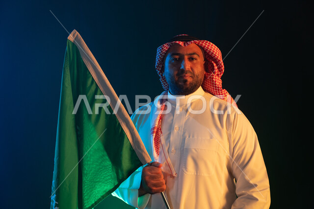 Looking at the camera and standing confidently, gestures of pride on Saudi Flag Day, March 11, showing feelings of pride in the homeland, commemoration of National Day on September 23, close-up portrait of a young Saudi Arabian Gulf man wearing a shemagh and traditional thobe, holding the Kingdom’s flag with respect and appreciation, black background