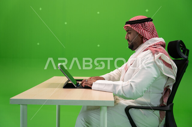 شركات ووظائف سعودية, مفهوم دمج العمل مع التقنية والتكنولوجيا, بورتريه من الجانب لرجل عربي سعودي خليجي يرتدي الزي السعودي التقليدي ينجز مهام العمل باستخدام جهاز التابليت, العمل في بيئة مريحة, خلفية خضراء, كروما
