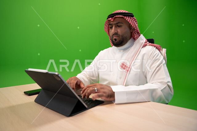 استخدام الأجهزة والتقنيات الحديثة, بورتريه مقرب لشاب عربي خليجي سعودي يرتدي الثوب السعودي التقليدي والشماغ يجلس في مكتبه, إنجاز مهام العمل عبر جهاز التابليت, متابعة وإدارة الأعمال الادارية, تصفح المواقع الالكترونية, خلفية خضراء, كروما