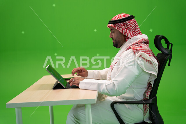 The concept of integrating work with technology and technology, a side-portrait of a Saudi Arabian Gulf man wearing traditional Saudi dress, performing work tasks using a tablet, Saudi companies and jobs, working in a comfortable environment, green backgr
