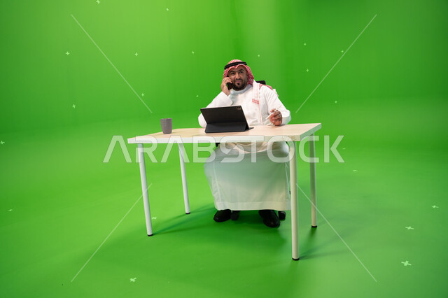 إجراء مكالمة هاتفية عبر الهاتف المحمول, بورتريه لموظف عربي سعودي خليجي يرتدي الثوب السعودي التقليدي والشماغ يقوم بالعمل داخل مكتبه, استخدام جهاز التابليت لإنجاز مهام العمل, إدارة شؤون الأعمال وتنظيمها, خلفية خضراء, كروما