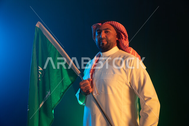 Commemorating the reunification of the nation, celebrating Saudi National Day on Rabi’ al-Awwal 8, portrait of a Saudi Gulf Arab man wearing a shemagh and traditional thobe holding the Kingdom’s flag with gestures of pride and pride, standing confidently on Saudi Flag Day March 11, black background with blue lighting