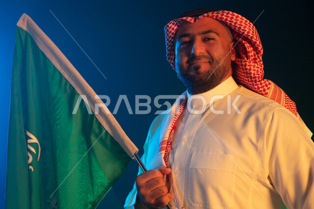 المشاركة في تنمية الوطن ورفع قدره, ذكرى اليوم الوطني السعودي في 8 ربيع الأول, بورتريه مقرب لرجل عربي سعودي خليجي مبتسم يرتدي الشماغ والثوب التقليدي يحمل علم المملكة بإيماءات السرور, الوقوف بثقة في يوم العلم السعودي 11 مارس, خلفية سوداء