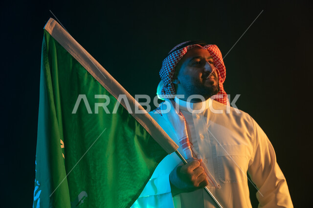 تخليد ذكرى إعادة توحيد الوطن, الاحتفال باليوم الوطني السعودي في 8 ربيع الأول, بورتريه مقرب لرجل عربي سعودي خليجي مبتسم يرتدي الشماغ والثوب التقليدي يحمل علم المملكة بإيماءات السرور, الوقوف بثقة في يوم العلم السعودي 11 مارس, خلفية سوداء