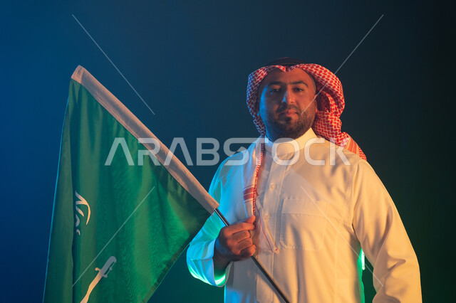 الوقوف بثقة والنظر إلى الكاميرا, إظهار مشاعر الاعتزاز بالوطن, ذكرى اليوم الوطني في 23 سبتمبر, بورتريه مقرب لرجل عربي سعودي خليجي يرتدي الشماغ السعودي والثوب يمسك علم المملكة باحترام, ايماءات السرور بيوم العلم السعودي 11 مارس, خلفية سوداء
