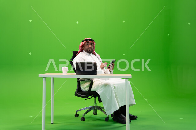 النظر إلى شيء ما, بورتريه لرجل عربي خليجي سعودي يرتدي الثوب السعودي والشماغ يستخدم الهاتف المحمول, مهن ووظائف سعودية, دمج التكنولوجيا مع الأعمال المكتبية, الجلوس في المكتب أمام جهاز التابليت, خلفية خضراء, كروما
