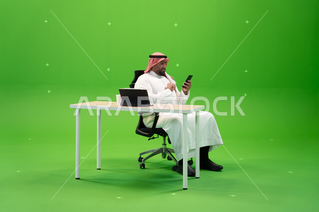 Sitting in the office and using modern applications and technologies, conversations and chats via social networking sites, a portrait of a Saudi Arabian Gulf citizen wearing traditional Saudi clothing holding a mobile phone, completing work tasks via the 