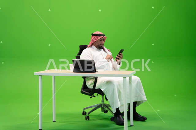 محادثات ودردشات عبر مواقع التواصل الاجتماعي, بورتريه لمواطن عربي سعودي خليجي يرتدي الزي التقليدي السعودي يمسك الهاتف المحمول, الجلوس في المكتب واستخدام التطبيقات والتقنيات الحديثة, إنجاز مهام العمل عبر الانترنت, خلفية خضراء, كروما