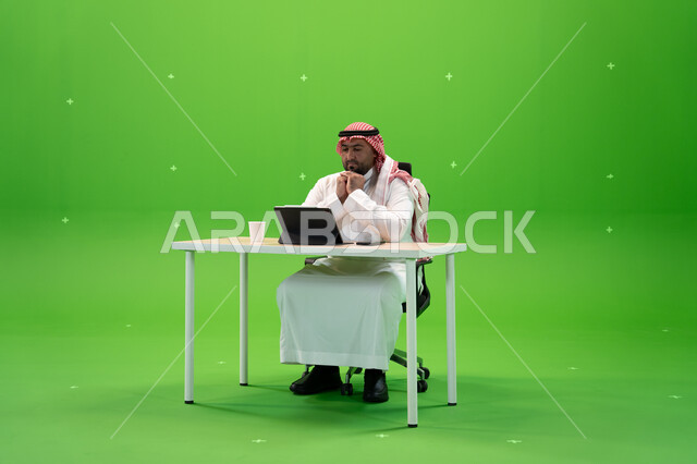 متابعة الشؤون الإدارية بإيماءات التفكير والتخطيط, النظر إلى جهاز التابليت وتطوير خطة العمل, إنجاز الأعمال المكتبية, بورتريه لموظف عربي سعودي خليجي يرتدي الثوب والشماغ التقليدي يقوم بالعمل داخل مكتبه, مهن ووظائف سعودية, خلفية خضراء, كروما