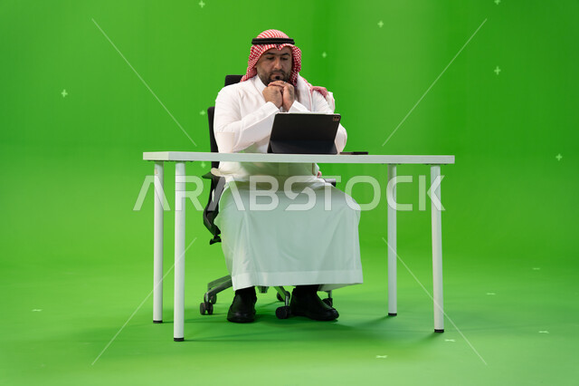 Looking at the tablet and developing a work plan, completing office work, portrait of a Saudi Gulf Arab employee wearing the traditional thobe and shemagh working inside his office, following up on administrative affairs with gestures of thinking and planning, Saudi professions and jobs, green background, chroma
