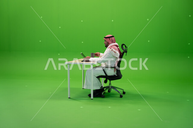 الاجتهاد في تطوير استراتيجة العمل, العمل في المجال المكتبي, بورتريه من الجانب لرجل أعمال عربي سعودي خليجي يرتدي الزي التقليدي يجلس في مكتبه أمام التابليت, استخدام التطبيقات الالكترونية في المكاتب والوظائف السعودية, الخلفية خضراء, كروما