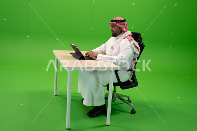 العمل في المجال المكتبي, الاجتهاد في تطوير استراتيجة العمل, بورتريه من الجانب لرجل أعمال عربي سعودي خليجي يرتدي الزي التقليدي يجلس في مكتبه أمام التابليت, استخدام التطبيقات الالكترونية في المكاتب والوظائف السعودية, الخلفية خضراء, كروما