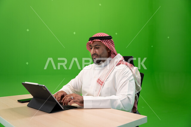 الأجهزة والتقنيات الحديثة المسستخدمة في المكاتب, بورتريه لشاب عربي خليجي سعودي يرتدي الثوب السعودي التقليدي والشماغ يجلس في مكتبه, إنجاز مهام العمل عبر جهاز التابليت, متابعة وإدارة الأعمال الادارية, تصفح المواقع الالكترونية, خلفية خضراء, كروما